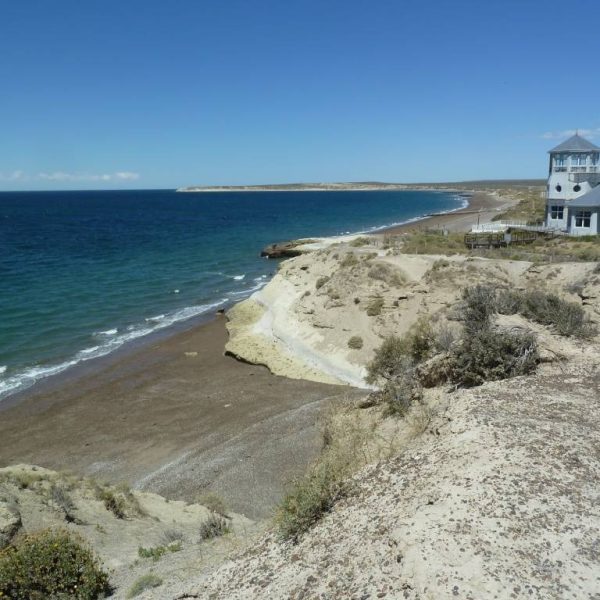 Das Bild zeigt eine Meeresküste. Das Meer liegt auf der linken Seite, aus der Mitte heraus ragt im Hintergrund eine Landzunge nach rechts ins Meer hinein. Auf der rechten Seite ist ein großes, weißes Haus mit einem grauen Dach zu erkennen, welches im hinteren Teil eine Art Aussichtsturm hat. Die Landschaft besteht abwechselnd aus Strand und Felsen mit einigen wenigen Sträuchern darin.