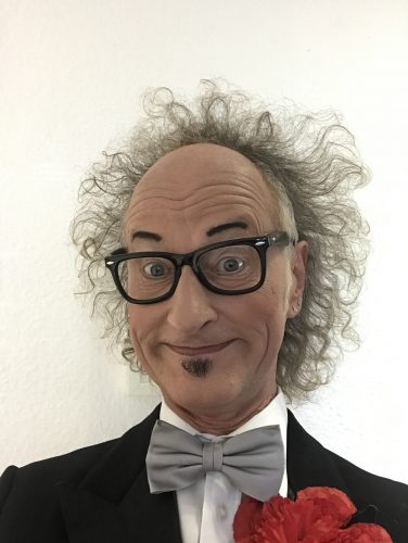 Das Bild zeigt einen Mann, der wie ein Clown aussieht. Seine Haare stehen wild in alle Richtungen vom Kopf ab, er hat eine schwarze Brille auf, trägt ein schwarzes Jackett, eine graue Fliege und hat eine rote Papier-Rose im Revers stecken. Der Mann guckt lustig in die Kamera.