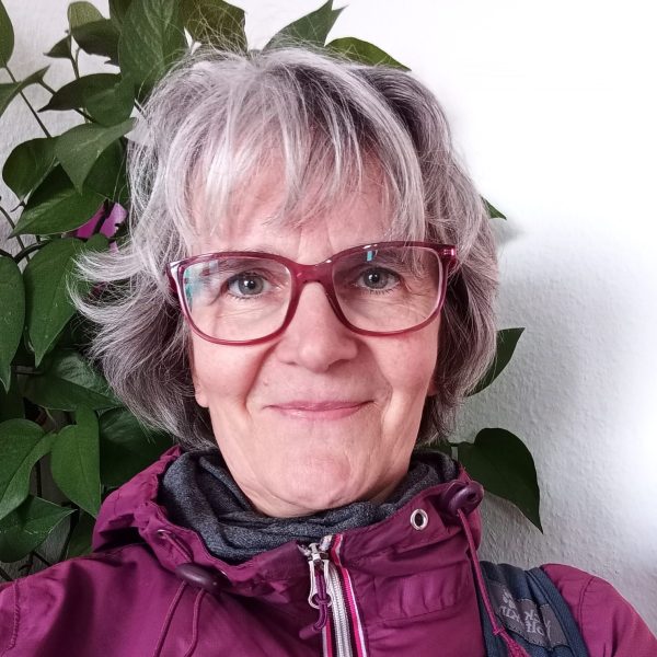 Das Bild zeigt eine Frau von mittlerem Alter mit grauen Haaren und einer größeren, rötlichen, Brille. Sie trägt eine lila Regenjacke und lächelt.