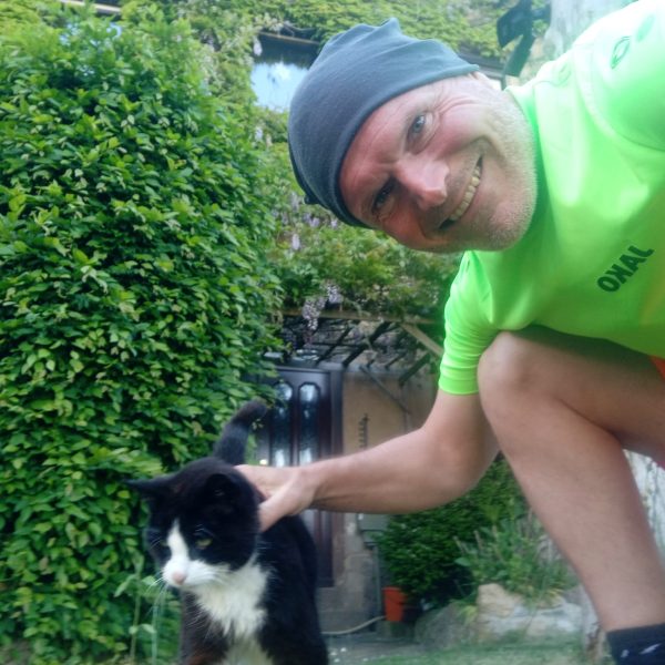 Das Bild zeigt einen Mann in einem gelben T-Shirt, einer orangenen Short und einer blauen Kopfbedeckung, der sich zu einer schwarzen Katze mit weißer Brust herunterbeugt und diese streichelt.